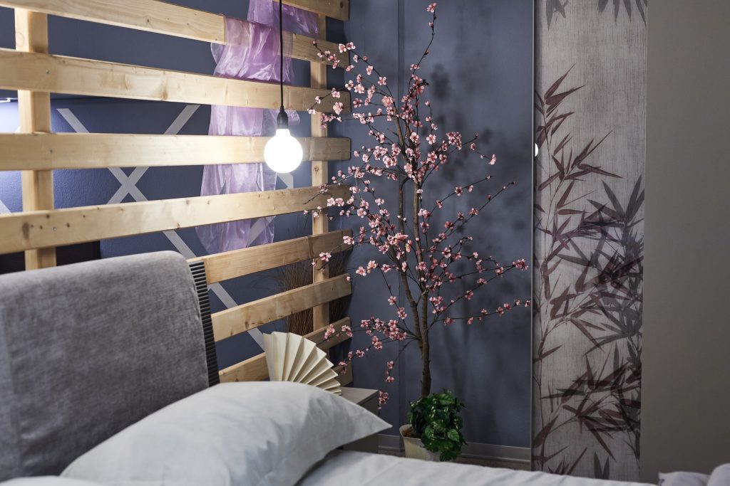 Letto matrimoniale con comodino e pianta ornamentale a fianco.