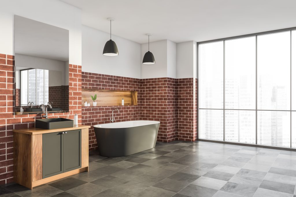 bagno dal design industrial, con mattonelle grigie a pavimento e rivestimenti in mattoni rossi. ampia finestra, lavabo in appoggio su mobile in legno con sportelli laccati grigi, ampio specchio, vasca freestanding in creamica laccata grigia