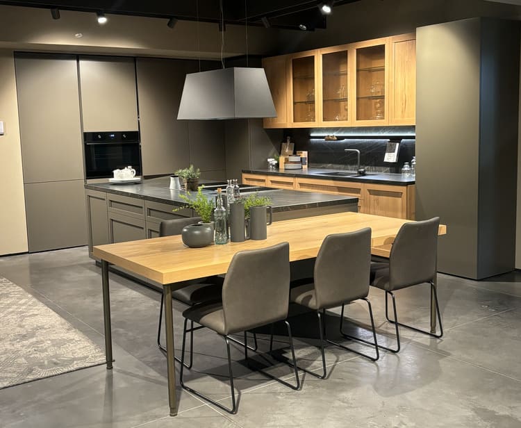Inquadratura panoramica di una cucina moderna con isola centrale e zona pranzo. In primo piano c’è un tavolo con piano in legno e quattro sedie grigie imbottite. Dietro, una grande isola centrale da cucina con piano di lavoro scuro e cappa di design sospesa. Sul fondo, una parete attrezzata a L con armadi scuri alti, basi in legno e pensili a giorno. Un forno integrato è visibile a sinistra e un lavello a destra. Il pavimento è in cemento grigio. L’illuminazione è costituita da faretti a binario.