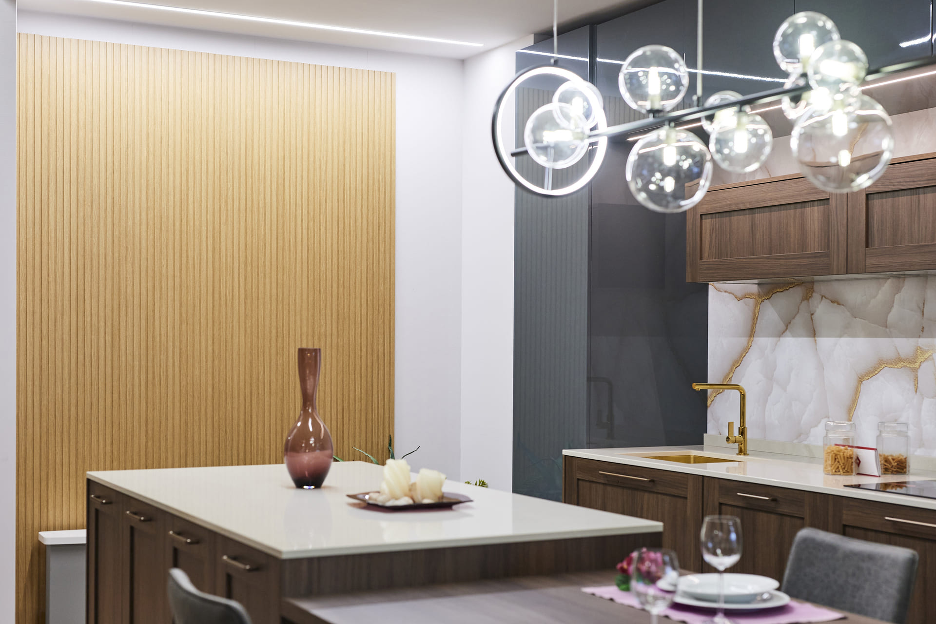Dettaglio di una cucina moderna con isola centrale in legno scuro e top bianco Inquadratura ravvicinata di una cucina moderna con isola centrale in legno scuro e top bianco. In alto c’è un lampadario a sospensione e sulla sinistra una parete rivestita da pannelli in legno effetto cannettato. A destra c’è una zona lavello con rubinetto e lavello dorati e un paraschizzi in marmo bianco con venature dorate. In primo piano a destra, una postazione da pranzo.